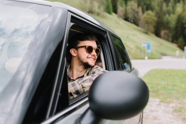 Handsome man in good mood drives a car and enjoying the fine weather on vacation. Smiling traveler wearing trendy sunglasses looking away on amazing nature view during trip around Europe..